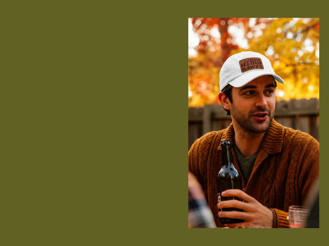 A man wearing a white ham radio baseball cap with a brown patch, ham radio and K4WYZ callsign on it.He has a brown cable-knit sweater, holding a dark bottle outdoors against a warm autumn background.