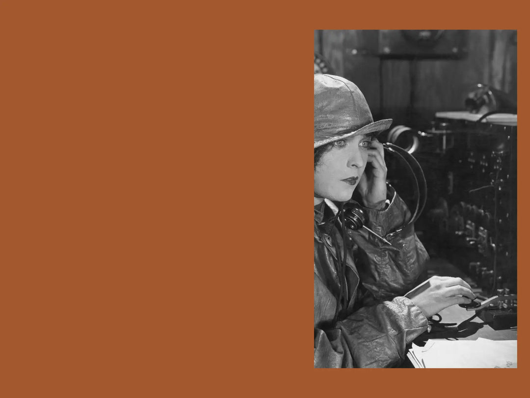 A black and white historical photograph of a woman in a leather jacket and hat speaking into a vintage radio transmitter, and send a morse code set against a solid terracotta orange background.