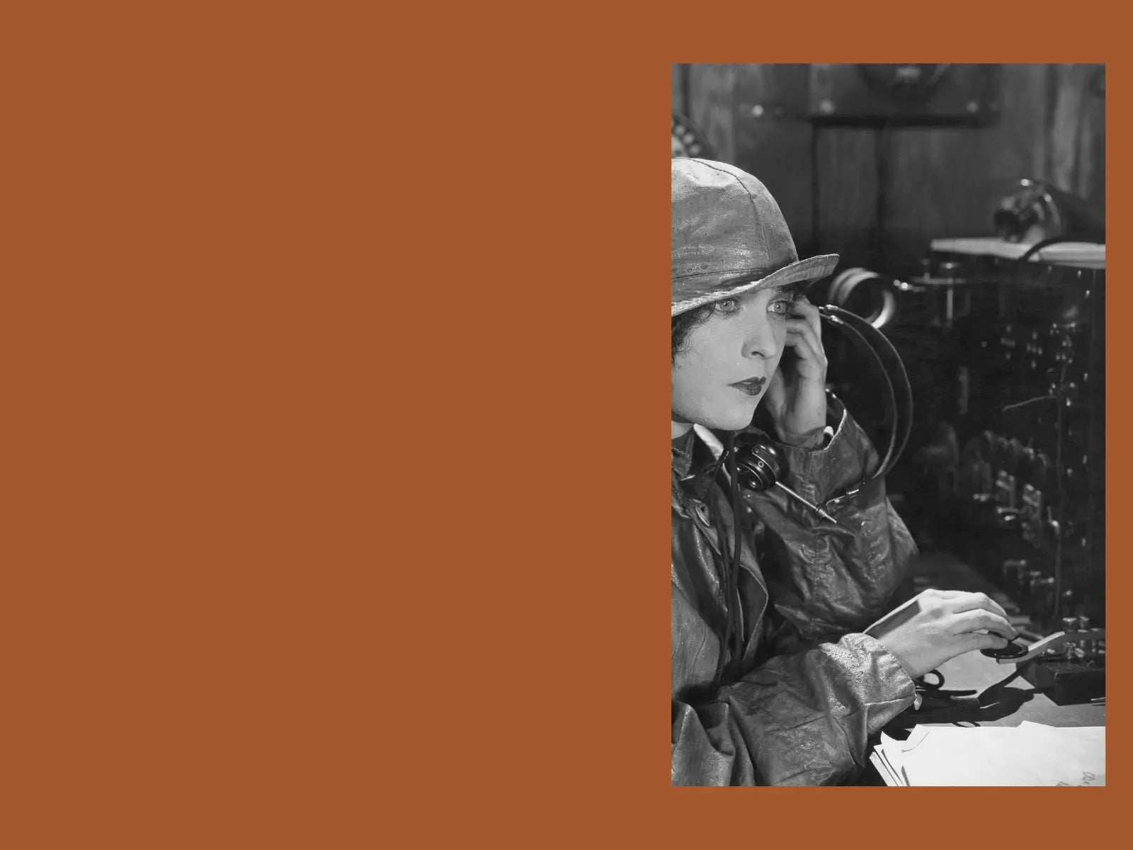 A black and white historical photograph of a woman in a leather jacket and hat speaking into a vintage radio transmitter, and send a morse code set against a solid terracotta orange background.