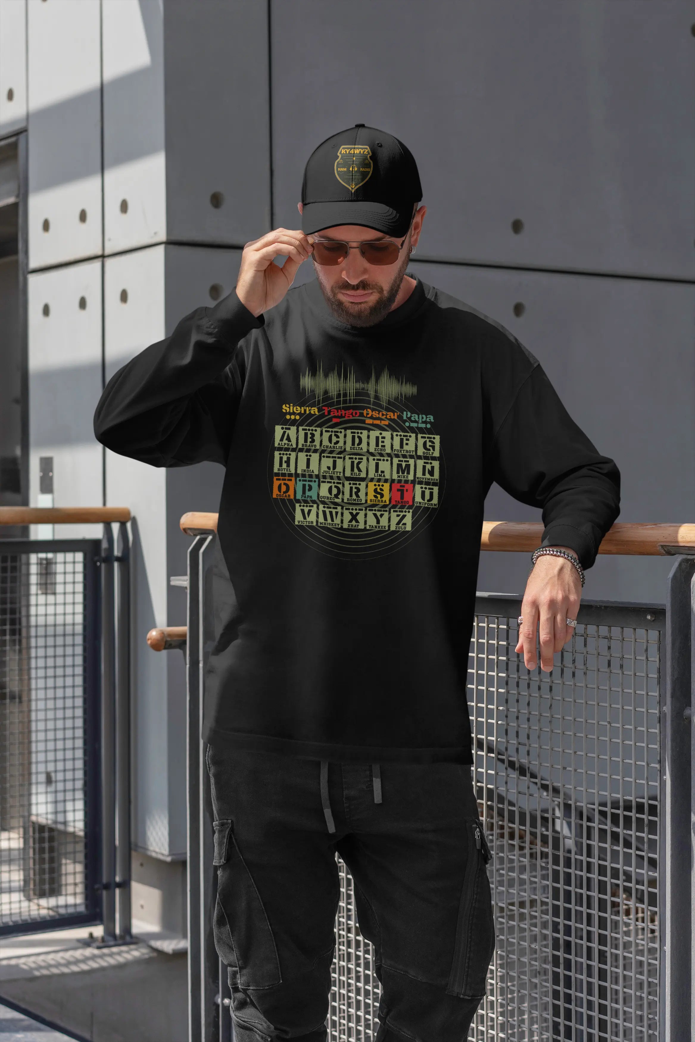 A man wearing a black long-sleeve graphic t-shirt, black cargo pants stands outdoors in front of a gray metal wall and railing. The shirt features a colorful phonetic alphabet and morse code graphic with the words 'Sierra', 'Tango', 'Oscar, and 'Papa'