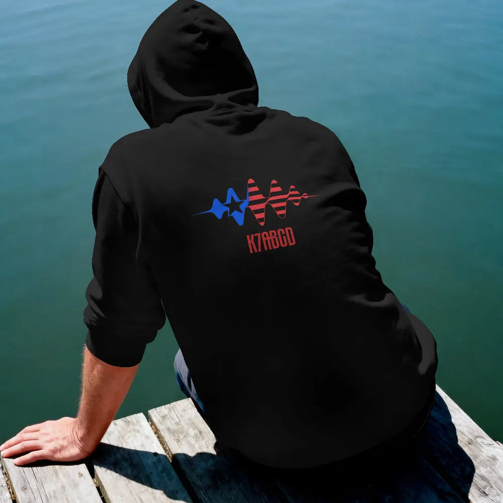 A person sitting on a wooden dock by the water, viewed from behind, wearing a black hoodie with a blue and red signal wave graphic and the ham radio callsign 'K7ABCD'' on the back.