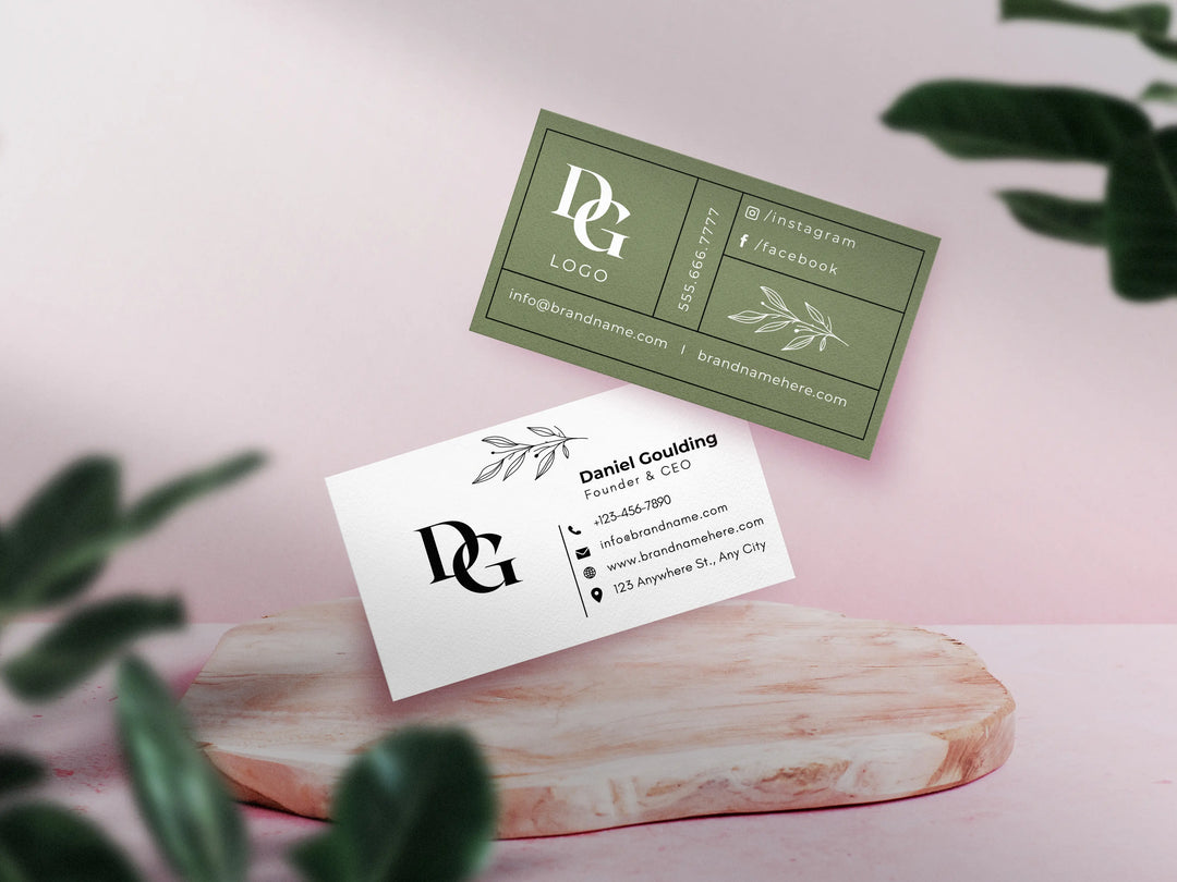 Two business cards with a logo on a pink surface with green leaves.
