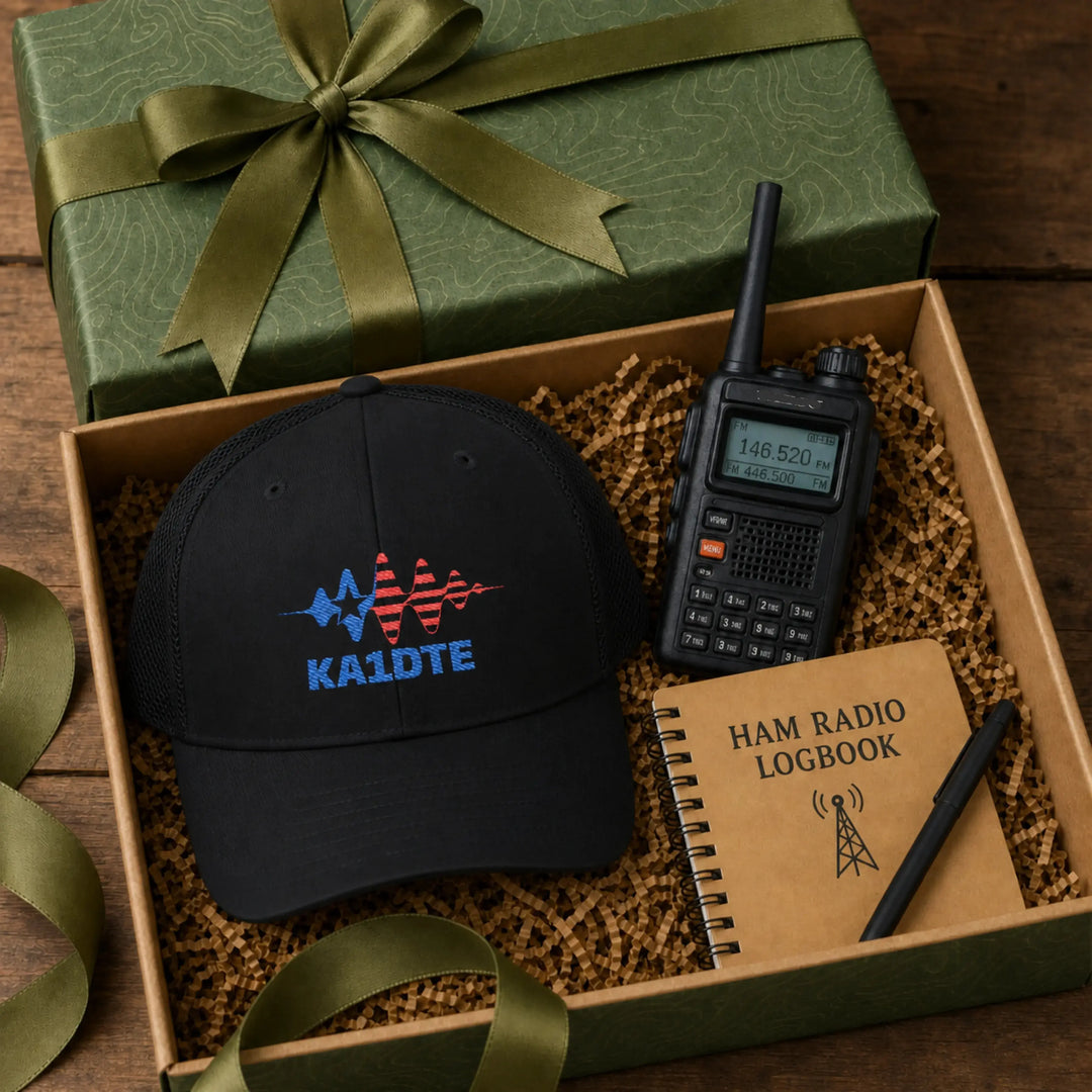 A gift set arranged in an open box with a green presentation box and ribbon, a black American flag signal wave ham radio callsign cap with a KA1DTE callsign, a black walkie-talkie, and a tan ham radio logbook on brown paper filler.