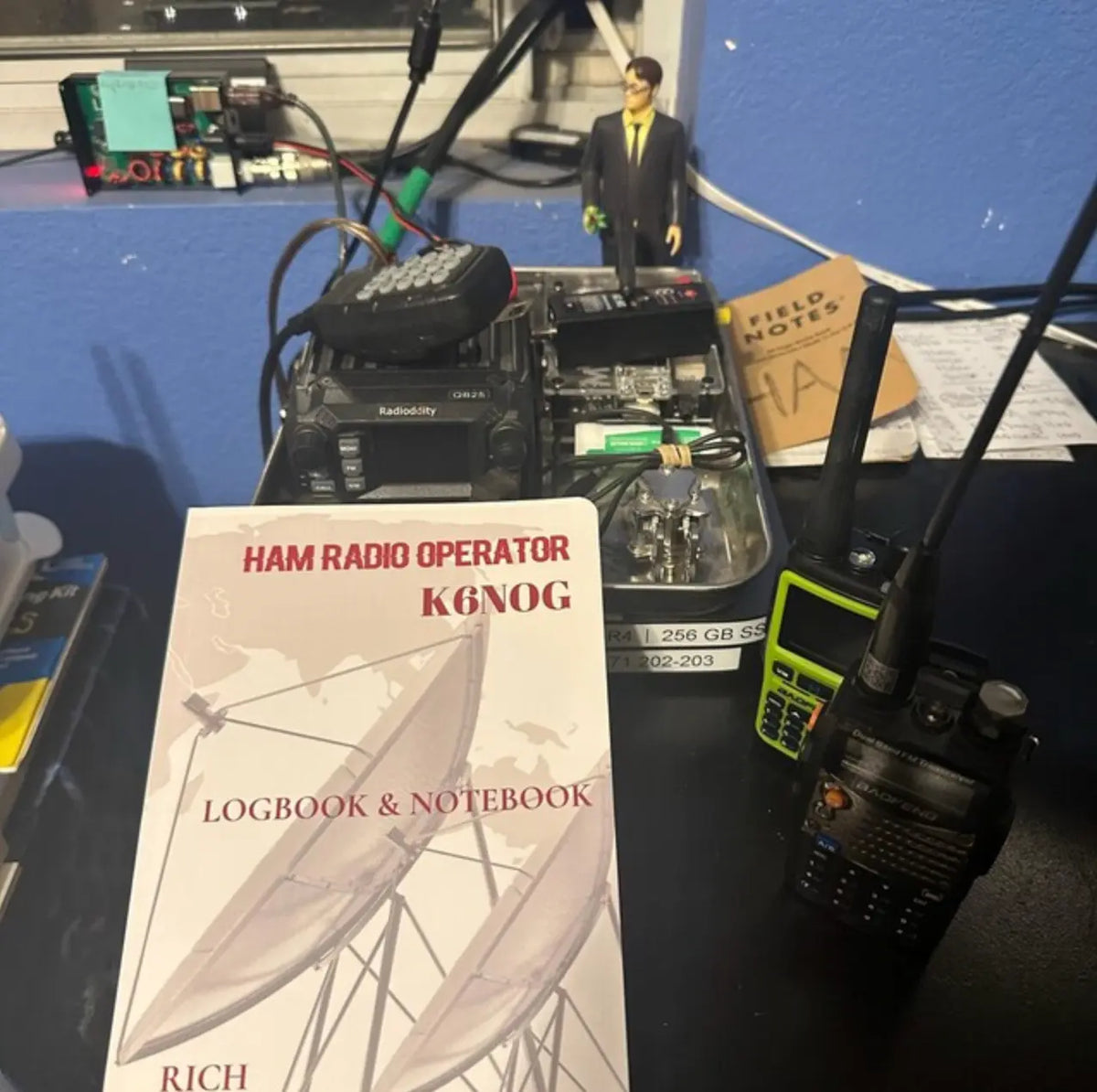 A Ham Radio Operator  logbook and notebook cover is displayed in the foreground on a desk, with walkie-talkies and electronic equipment in the background against a blue wall.