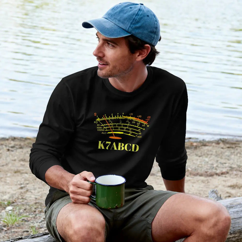 A man sitting outdoors by the water wearing a black long-sleeve graphic t-shirt with a yellow and orange S-meter graphic and the ham radio callsign 'K7ABCD', a blue baseball cap, and green shorts, holding a green enamel mug.