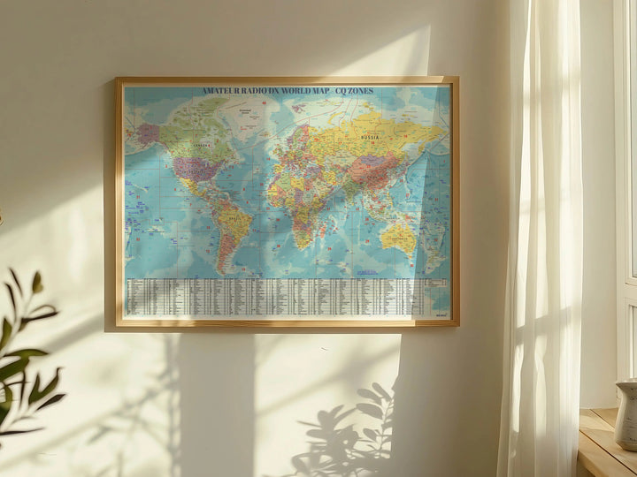 A framed amateur radio world map poster hanging on a sunlit cream wall, with warm sunlight and window shadows cast across the room. The map shows Africa, Europe, Asia, and South America in muted pastel colors, with a title bar at the top and a CQ &amp; ITU Zones on the bottom.