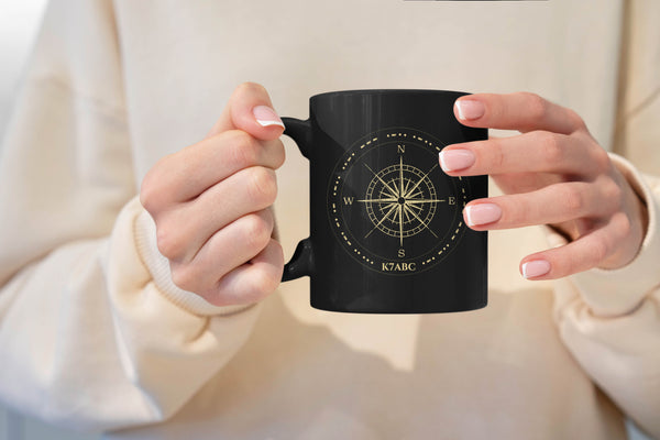 A person holding a black ceramic mug with a gold compass rose design and the text 'K7ABC' on the front, wearing a cream-colored sweatshirt.